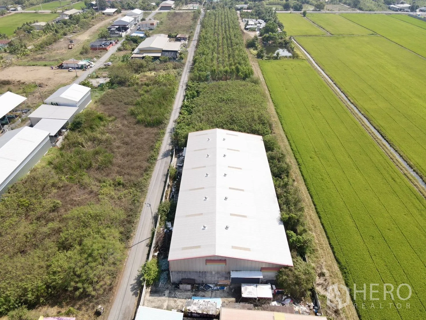 仓库 for sale in Lam Luk Ka, Pathum Thani - 俯瞰带屋脊通风口的仓库，旁有乡道、农田及邻近建筑。