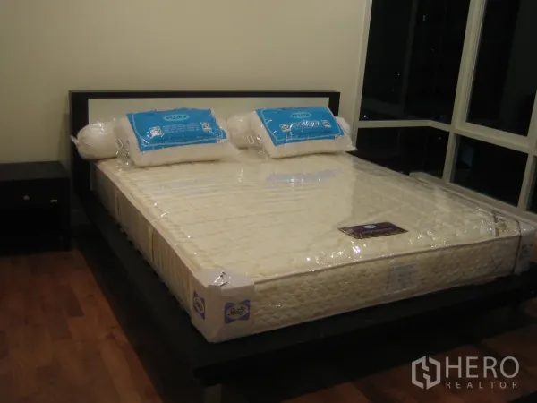 Condominium for rent or sale in Watthana, Bangkok - Master bedroom with a new mattress on a platform bed, bedside table, wood floors, and large corner windows.