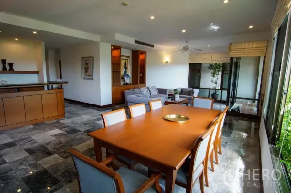 Apartment for rent in Watthana, Bangkok - Open-plan dining and living room with marble floors, wooden table for eight, built-in cabinetry, and large windows leading to a balcony.