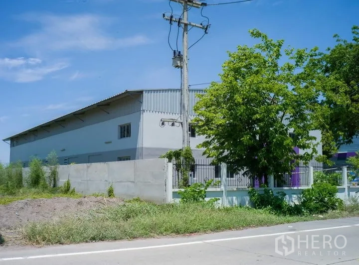Factory for rent in Pathum Thani - Industrial building set behind a fence along a main road with mature trees and power pole.
