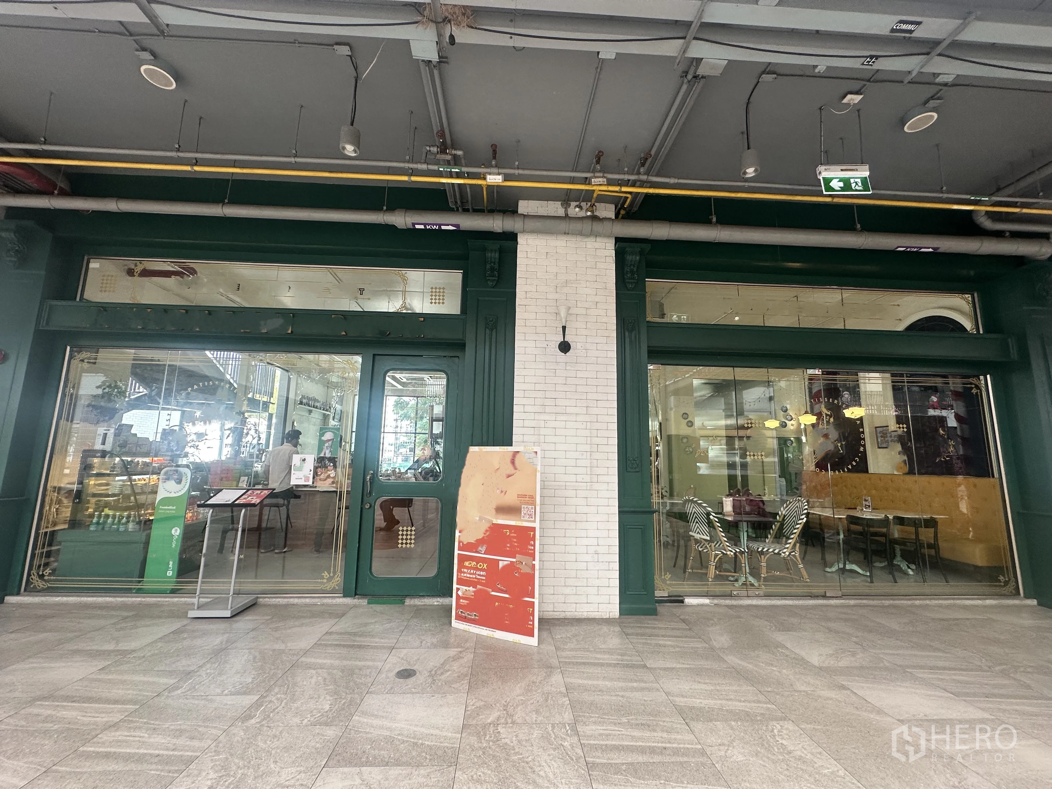 零售空间 for sale in Khlong Toei, Bangkok - 绿色框架的玻璃店面，双开门与大面积展示橱窗，位于有顶商业走廊的抛光石地面前。