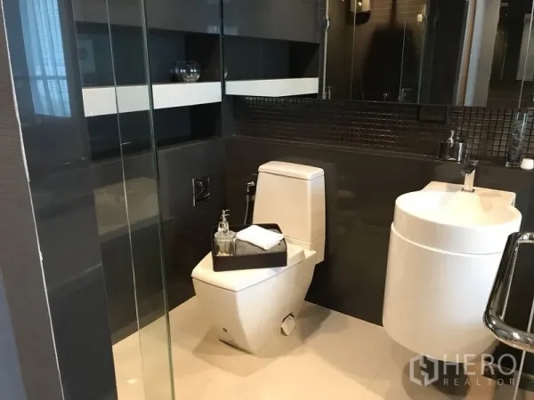 Condominium for rent in Sathon, Bangkok - Modern bathroom with glass shower, square toilet and round basin set against dark accent tiles.