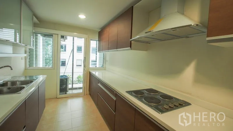 Condominium for rent in Khlong San, Bangkok - Galley kitchen view showing electric cooktop, range hood, long counters, and balcony access.