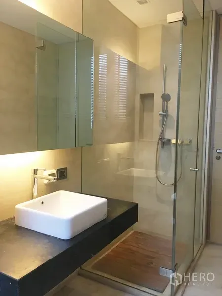 Condominium for rent in Bang Rak, Bangkok - Modern bathroom featuring a glass-enclosed shower, square vessel sink, and warm backlit mirror.