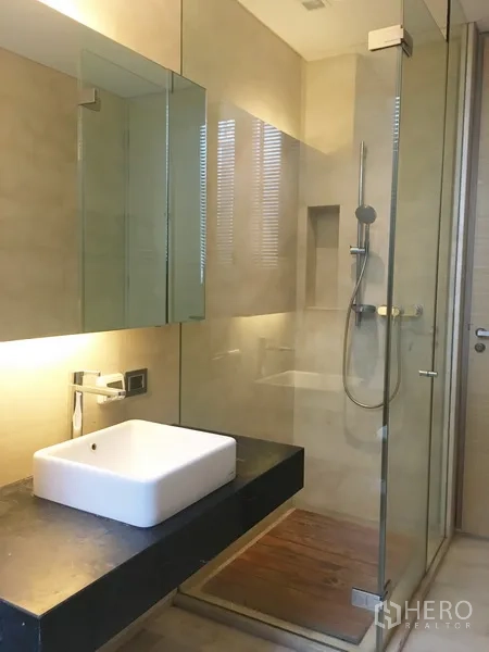 Condominium for rent in Bang Rak, Bangkok - Modern bathroom featuring a glass-enclosed shower, square vessel sink, and warm backlit mirror.