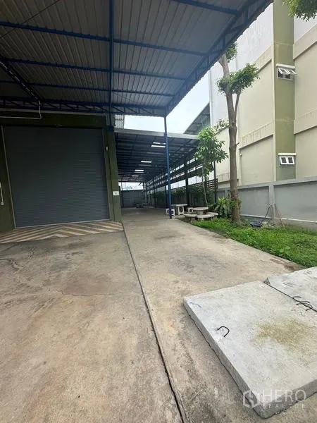 Warehouse for rent or sale in Lam Luk Ka, Pathum Thani - Covered driveway with roller shutter loading door and small landscaped strip.