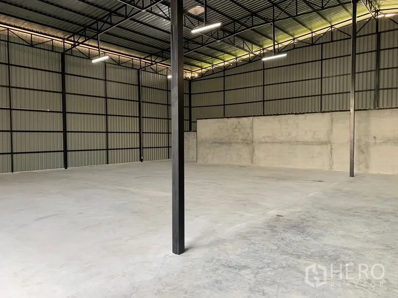 Warehouse for rent in Khan Na Yao, Bangkok - Wide view of the warehouse showing metal cladding, steel columns, bright lighting, and open concrete slab.