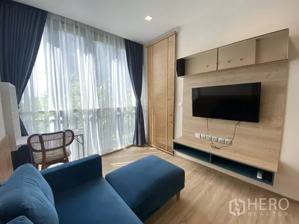 Condominium for rent or sale in Watthana, Bangkok - Bright living room with floor-to-ceiling curtains, blue sofa, wood floors and a wall-mounted TV unit.