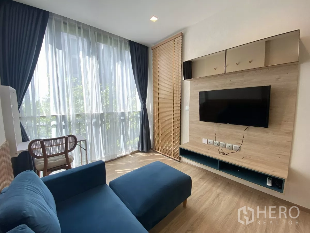 Condominium for rent or sale in Watthana, Bangkok - Bright living room with floor-to-ceiling curtains, blue sofa, wood floors and a wall-mounted TV unit.