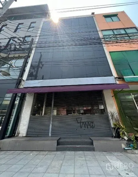 店屋 出租在Watthana, Bangkok - 临街多层商铺，黑色玻璃立面，紫色雨棚与卷闸门。