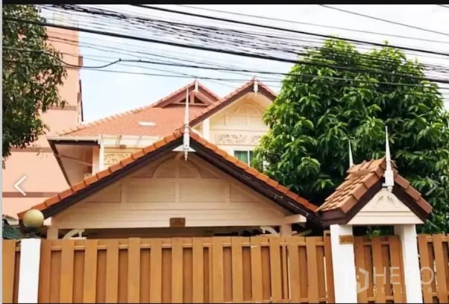 Detached House for sale in Saphan Sung, Bangkok - Street-facing view of a detached house with terracotta gable roof, tall wooden fence and mature trees in Saphan Sung.