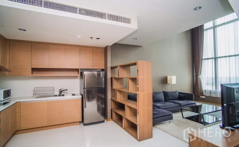 Condominium for rent or sale in Khlong Toei, Bangkok - Open-plan kitchen with wood cabinetry and stainless fridge next to living area.