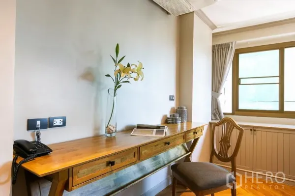 Apartment for rent in Watthana, Bangkok - Sunny study nook with a long wooden desk, vase and chair beside a curtained window.