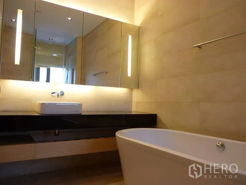 Condominium for rent in Bang Rak, Bangkok - Bathroom vanity with long black countertop, illuminated mirrors and a deep soaking tub.