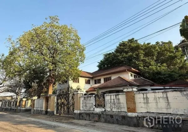 Detached House for sale in Sai Mai, Bangkok - Two-storey detached house in Sai Mai, Bangkok with red-tile roof, mature trees and an ornate wrought-iron gate on a quiet street.