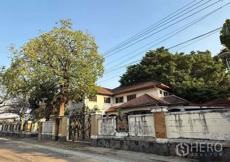 Detached House for sale in Sai Mai, Bangkok - Two-storey detached house in Sai Mai, Bangkok with red-tile roof, mature trees and an ornate wrought-iron gate on a quiet street.