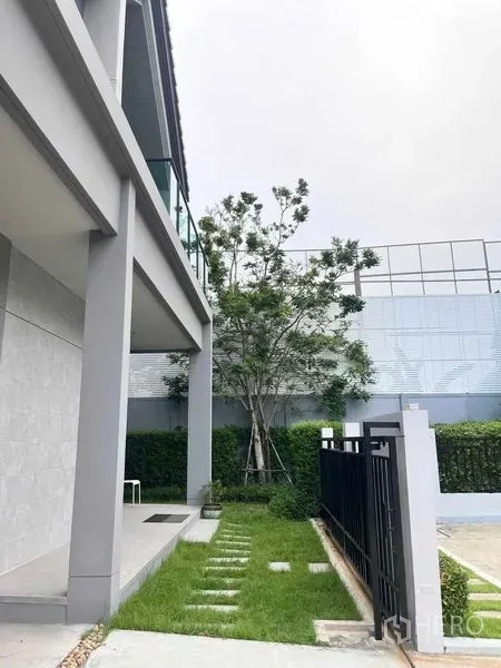 Detached House for sale in Bang Kruai, Nonthaburi - Side yard with green lawn, stepping stones, tall tree and privacy fence beside a covered terrace.