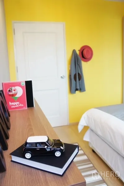 Condominium for rent in Phaya Thai, Bangkok - Bedroom corner with yellow accent wall, dresser, decorative items and bed edge by a white door.