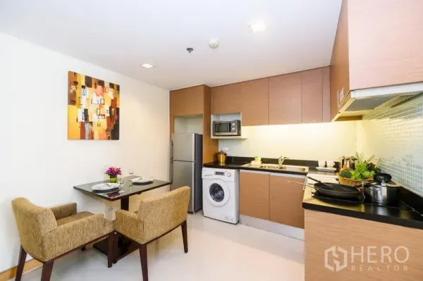 Condominium for rent in Khlong Toei, Bangkok - Modern open kitchen with wood cabinets, washing machine, microwave, fridge and a two-seat dining table.