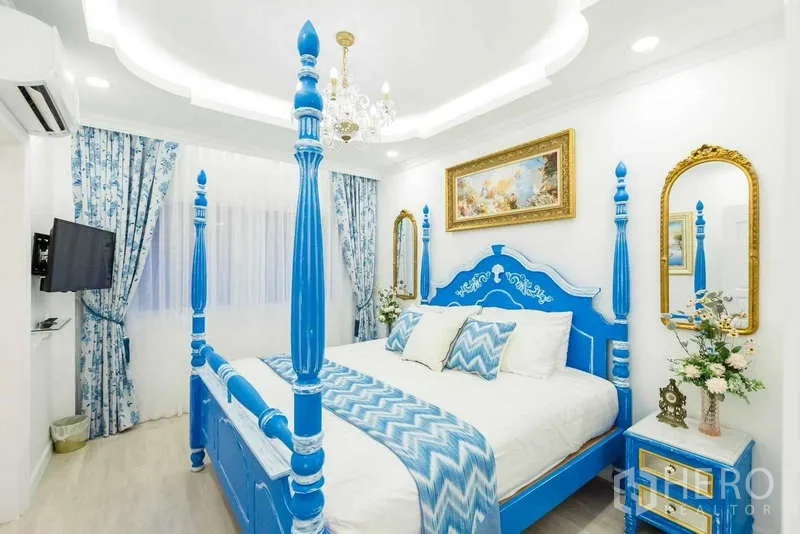 Townhouse for rent in Lat Phrao, Bangkok - Blue four-poster bedroom with chandelier, patterned curtains and gold-trimmed mirrors.