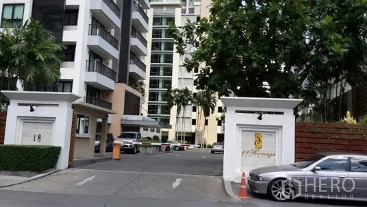 公寓 出租或出售在Watthana, Bangkok - 现代公寓小区大门，车道、绿树与停放车辆。