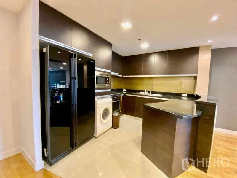 Condominium for rent or sale in Sathon, Bangkok - Corner kitchen with black granite counters, side-by-side fridge, microwave, and washer.