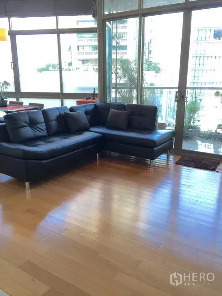 Condominium for rent in Khlong Toei, Bangkok - Corner living room with black L-shaped sofa, floor-to-ceiling windows and balcony showcasing city views.