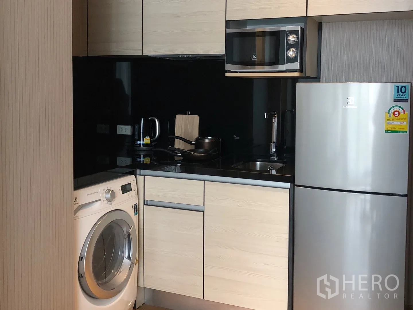 Condominium for rent in Khlong Toei, Bangkok - Compact modern kitchenette with black backsplash, microwave, fridge and front-load washer.