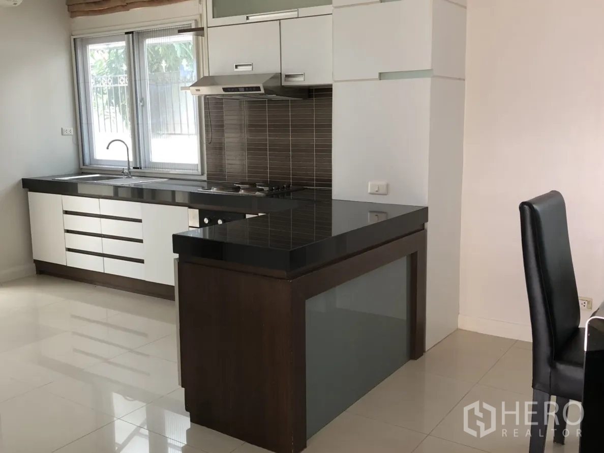 Detached House for rent or sale in Lat Phrao, Bangkok - L-shaped kitchen with black granite counters, white cabinetry and a brown tile backsplash.
