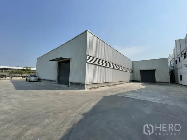Warehouse for rent in Mueang Samut Prakan, Samut Prakan - Modern metal-clad warehouse with two large roller shutter doors and a wide concrete yard for truck access under a clear sky.