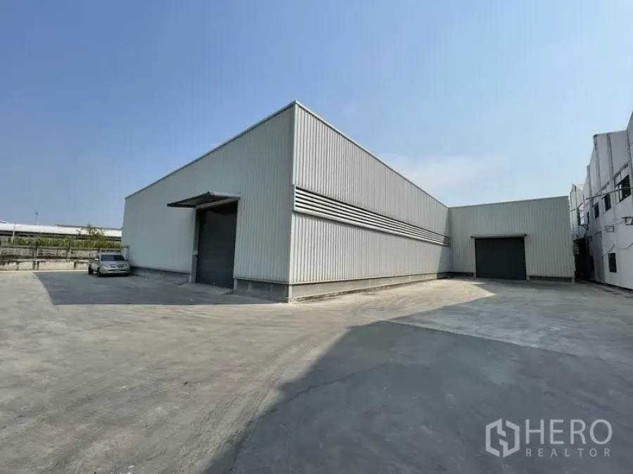 Warehouse for rent in Mueang Samut Prakan, Samut Prakan - Modern metal-clad warehouse with two large roller shutter doors and a wide concrete yard for truck access under a clear sky.