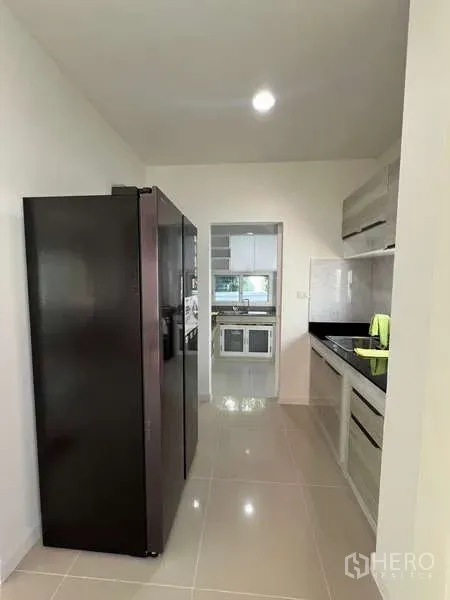 Detached House for rent in Bang Kapi, Bangkok - Galley kitchen with tall black fridge and built‑in cabinets leading to a Thai kitchen.