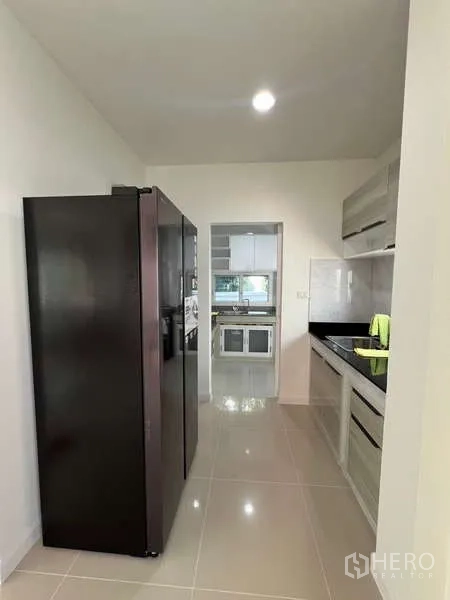 Detached House for rent in Bang Kapi, Bangkok - Galley kitchen with tall black fridge and built‑in cabinets leading to a Thai kitchen.