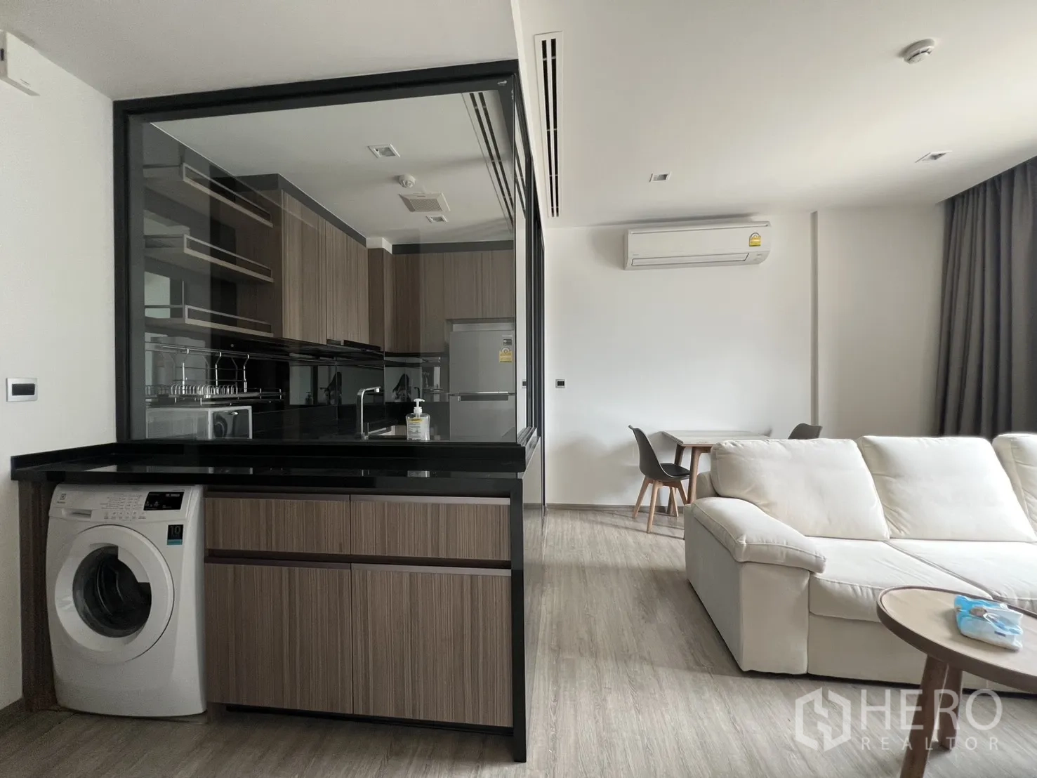 คอนโด for rent in Watthana, Bangkok - ครัวสมัยใหม่ท็อปสีดำหลังผนังกระจก มองออกไปยังโซฟาสีขาวและโต๊ะกินข้าวขนาดเล็ก.