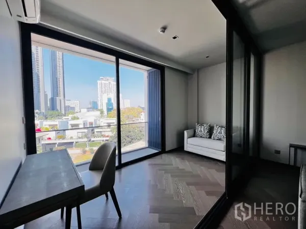 公寓 出售在Watthana, Bangkok - 明亮起居与书写区，落地推拉门通往阳台，城市天际线景观，鱼骨拼木地板、书桌与小沙发。