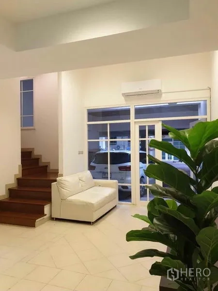 Townhouse for rent or sale in Chatuchak, Bangkok - Bright living area with white sofa, wood stairs, indoor plant and glass doors opening to a covered parking space.