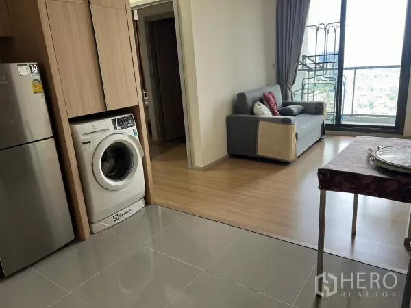公寓 出租或出售在Chatuchak, Bangkok - 开放式起居区，小型沙发连通阳台，旁侧为洗衣机和冰箱柜体。