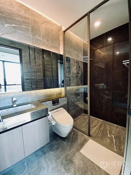 Condominium for rent in Khlong Toei, Bangkok - Modern gray-tiled bathroom with floating vanity, smart toilet, and glass-enclosed rain shower.