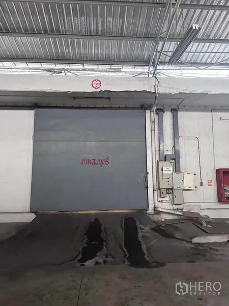 Warehouse for rent in Mueang Samut Prakan, Samut Prakan - Close-up of a steel roller shutter door with loading ramp and utility boxes.