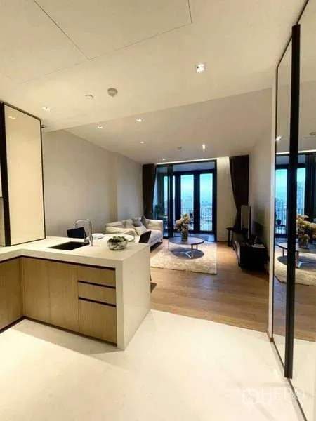 Condominium for rent or sale in Khlong Toei, Bangkok - Open-plan living area with modern kitchenette, wood floors, and balcony doors facing a city view.