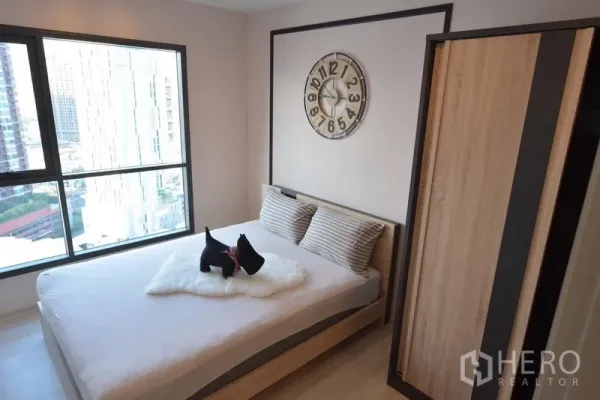 Condominium for sale in Khlong Toei, Bangkok - Cozy bedroom with large window, queen bed, wood wardrobe and wall clock, offering a city view.