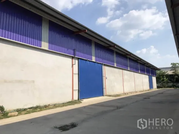 Warehouse for rent in Sai Noi, Nonthaburi - Side exterior of a long warehouse with purple metal cladding, a blue sliding steel door and a concrete driveway under a clear sky.