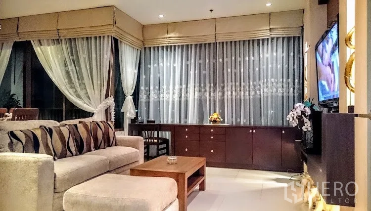 Condominium for rent in Khlong Toei, Bangkok - Warm living room with beige sofa, built-in cabinets and floor-to-ceiling windows with sheer curtains.