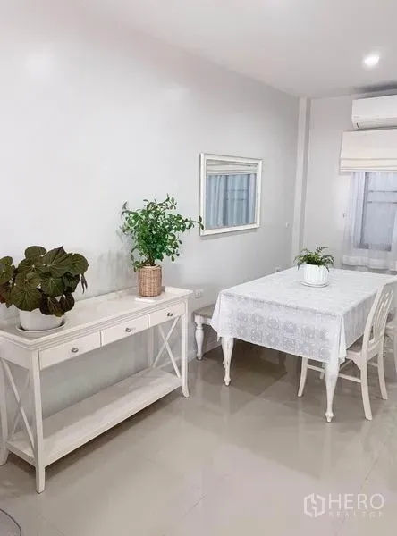 联排别墅 出租在Prawet, Bangkok - 温馨用餐角，白色餐桌、边柜、绿植与百叶窗。