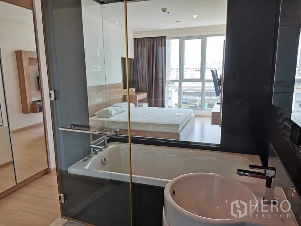 Condominium for rent or sale in Khlong Toei, Bangkok - Glass-walled bathroom with a bathtub looking into a bright bedroom with large windows and city outlook.