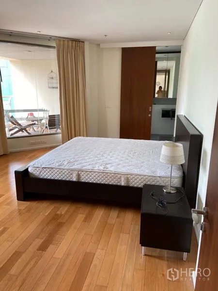 Condominium for rent in Khlong Toei, Bangkok - Spacious master bedroom with wooden floors, bedside table, and access to a balcony.