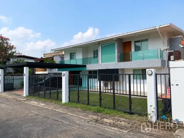 Home Office for rent in Chatuchak, Bangkok - Two-storey modern home office with glass balconies, front lawn, black metal fence, and a covered carport on a quiet street.