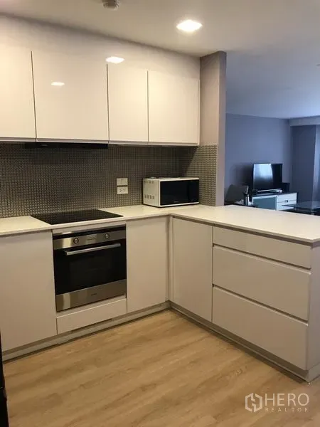 Condominium for rent in Bang Rak, Bangkok - L-shaped kitchen with glossy cabinets, built-in oven, electric cooktop and microwave facing the living area.
