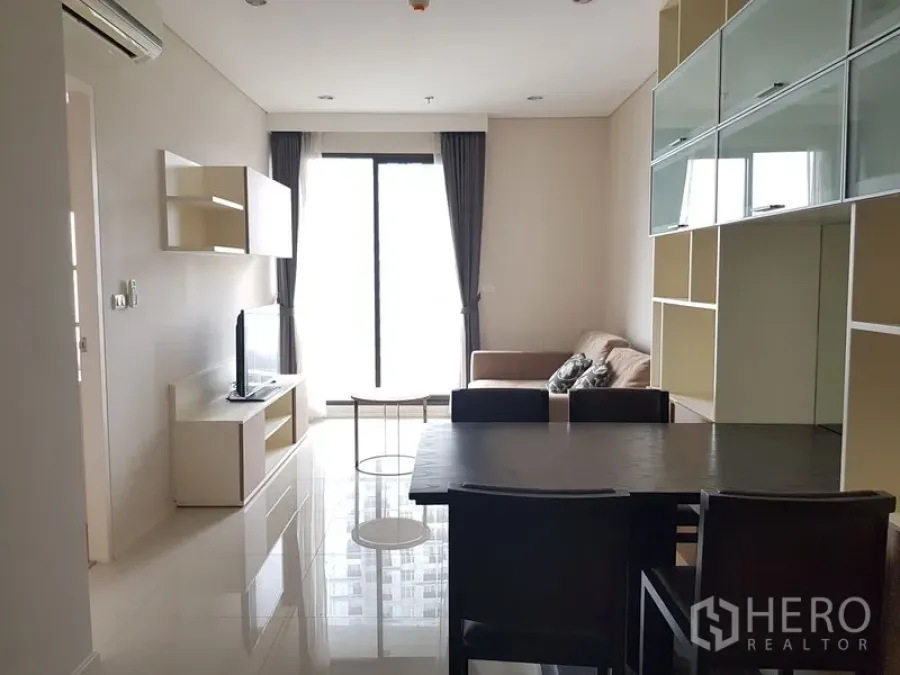 Condominium for rent in Ratchathewi, Bangkok - Bright living and dining area in a Ratchathewi condo with sofa, TV console, dark dining set, built-in shelving and floor‑to‑ceiling window to the balcony.