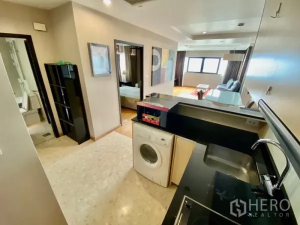 Condominium for rent or sale in Sathon, Bangkok - Open-plan kitchen with black countertop and washing machine leading to the living area and bedroom in a Sathorn condo.